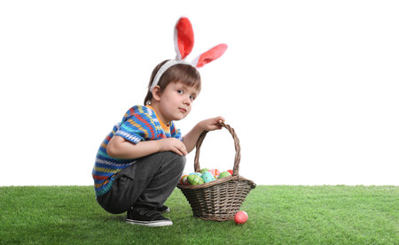 Cute little boy wearing bunny ears with basket full of dyed Easter eggs on green grassの写真素材