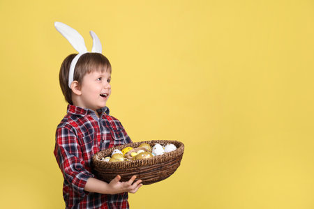 Cute little boy wearing bunny ears with basket full of dyed Easter eggs on yellow background