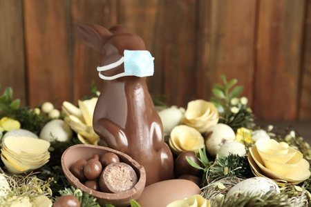 Chocolate bunny with protective mask, eggs and wreath against wooden background, closeup. Easter holiday during COVID-19 quarantineの写真素材