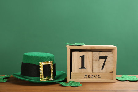 Leprechaun hat, clover leaves and block calendar on wooden table against green background, space for text. St Patrick's Day celebrationの写真素材
