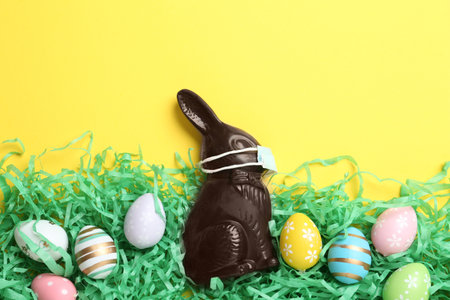 Chocolate bunny with protective mask and eggs on yellow background, flat lay. Easter holiday during COVID-19 quarantineの写真素材