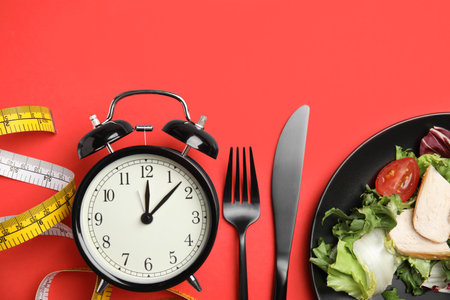 Plate of tasty salad, cutlery, alarm clock and measuring tape on red background, flat lay. Nutrition regimenの写真素材