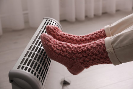 Woman warming feet on electric heater at home, closeup