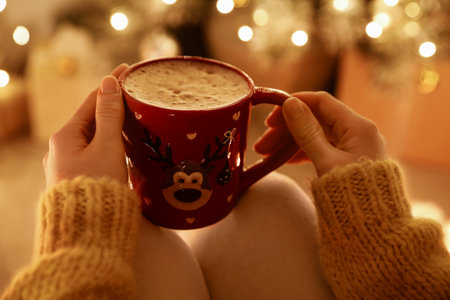 Woman with cup of drink and blurred Christmas lights on background, closeupの写真素材