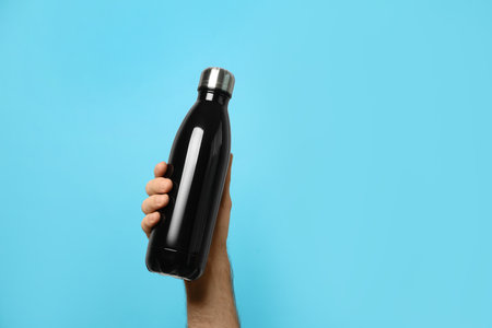 Man holding black flask bottle on light blue background, closeup. Space for textの写真素材