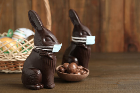 Chocolate bunnies with protective masks and eggs on wooden table, space for text. Easter holiday...