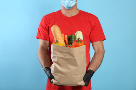 Courier in medical mask holding paper bag with food on light blue background, closeup. Delivery service during quarantine due to Covid-19 outbreakの写真素材