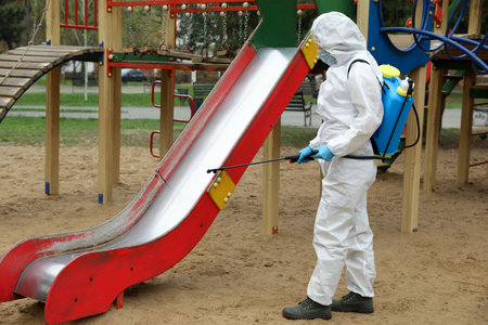 Woman wearing chemical protective suit with disinfectant sprayer on playground. Preventive measure during coronavirus pandemicの写真素材
