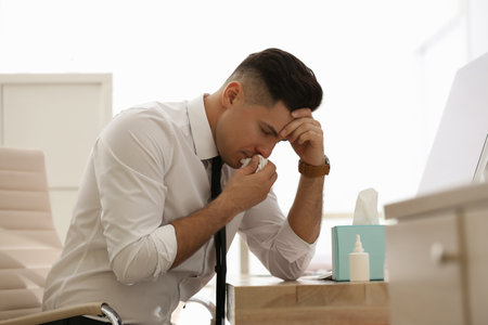 Ill businessman at table with nasal spray and box of paper tissues in officeの写真素材