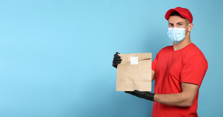 Courier in medical mask holding paper bag with takeaway food on light blue background, space for text. Delivery service during quarantine due to Covid-19 outbreakの写真素材