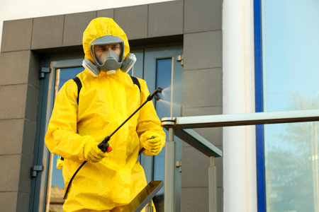 Person in hazmat suit with disinfectant sprayer cleaning metal railing on city street. Surface treatment during coronavirus pandemicの写真素材