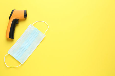 Infrared thermometer and medical mask on yellow background, flat lay with space for text. Checking...