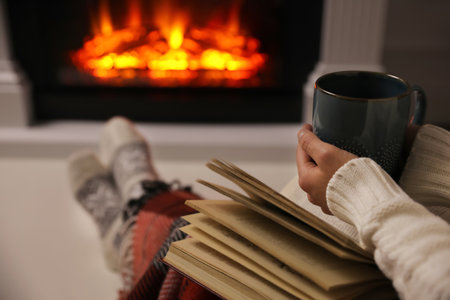 Woman with cup of hot drink and book resting near fireplace at home, closeupの写真素材