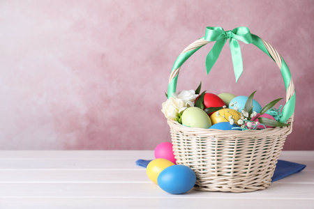 Wicker basket with bright painted Easter eggs and flowers on white wooden table against pink background. Space for textの写真素材