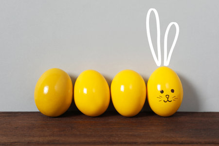 One egg with drawn face and ears as Easter bunny among others on wooden table against light gray backgroundの写真素材