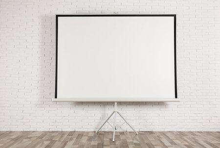 Blank projection screen near white brick wall indoors. Space for designの写真素材