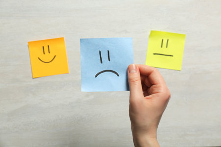 Woman holding sticker with sad face on light background, closeupの写真素材