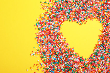 Heart shaped frame of sprinkles on yellow background, flat lay with space for text. Confectionery decorの写真素材