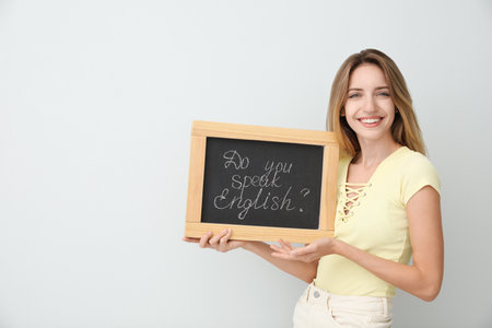 Young female teacher holding chalkboard with words DO YOU SPEAK ENGLISH? on light background. Space for textの写真素材