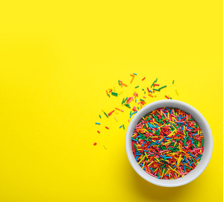 Colorful sprinkles in bowl on yellow background, top view with space for text. Confectionery decorの写真素材