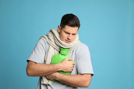 Ill man with hot water bottle suffering from cold on light blue backgroundの写真素材