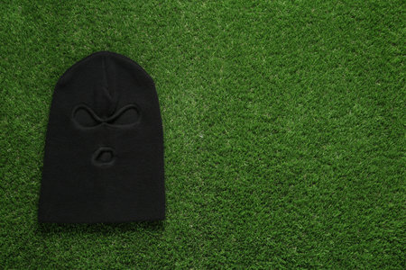 Black knitted balaclava on green grass, top view. Space for text