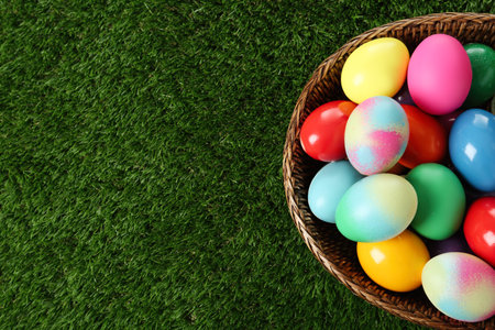 Wicker basket with bright painted Easter eggs on green grass, top view. Space for text