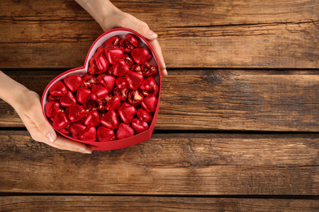 Woman holding box with heart shaped chocolate candies on wooden table, top view. Space for textの写真素材
