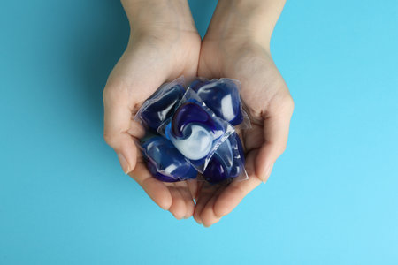 Woman holding laundry capsules on light blue background, top view