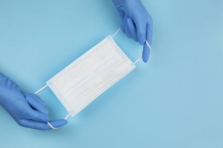 Doctor in medical gloves holding protective mask on light blue background, closeup. Space for text