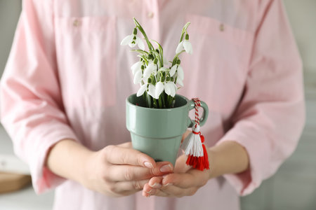 Woman holding pot with beautiful snowdrops and traditional martisor indoors, closeup. Symbol of first spring dayの写真素材