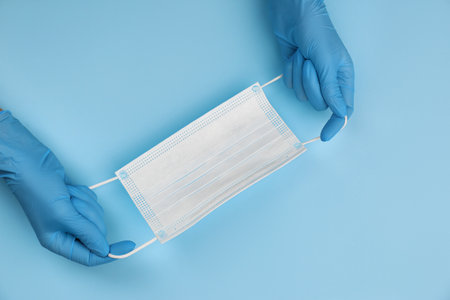 Doctor in medical gloves holding protective mask on light blue background, closeup. Space for textの写真素材