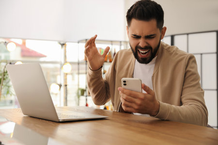 Emotional young man with smartphone at table in office. online hate conceptの写真素材