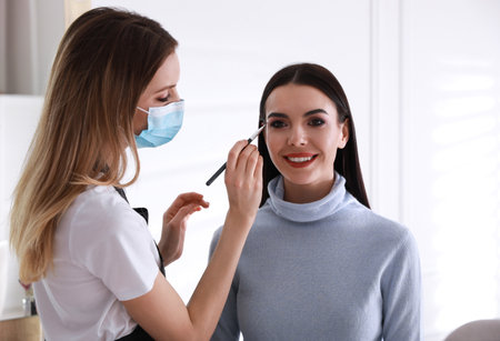 Makeup artist in protective mask working with woman indoors. Preventive measures during COVID-19...