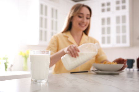 Woman with gallon bottle and breakfast cereal at white marble table in kitchen, focus on glass of milkの写真素材