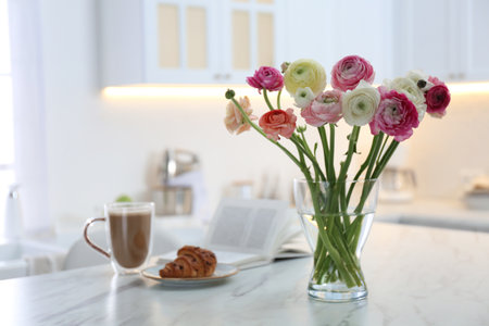 Beautiful fresh ranunculus flowers near cup of coffee, croissant and book on table in kitchenの写真素材