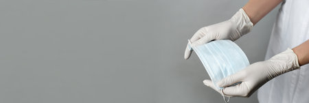 Doctor in medical gloves holding protective mask on light gray background, closeup. Banner design with space for textの写真素材