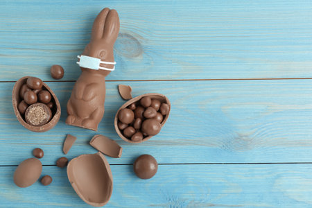 Chocolate bunny with protective mask, eggs and space for text on light blue wooden table, flat lay. Easter holiday during COVID-19 quarantineの写真素材