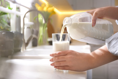 Young woman pouring milk from gallon bottle into glass at white countertop in kitchen, closeupの写真素材