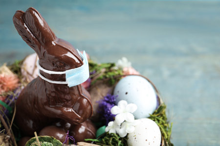 Chocolate bunny with protective mask, eggs, wreath and space for text on light blue wooden table, closeup. Easter holiday during COVID-19 quarantineの写真素材