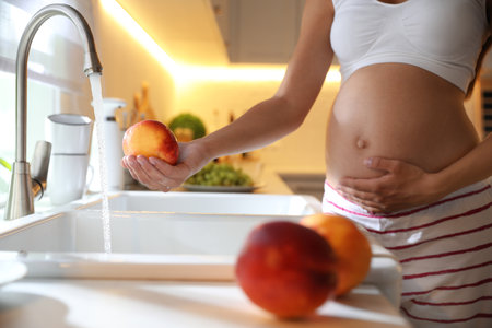 Young pregnant woman washing fresh sweet peach in kitchen, closeup. Taking care of baby healthの写真素材