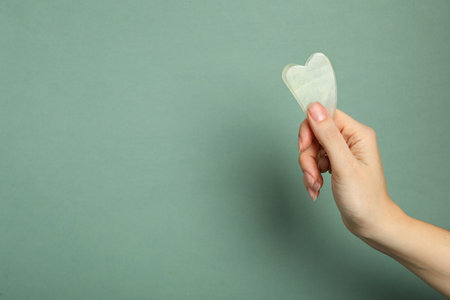Woman with jade gua sha tool on gray green background, closeup. Space for textの写真素材