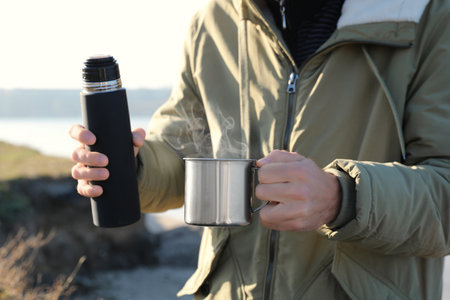 Man holding thermo and aluminum mug with hot drink outdoors, closeup