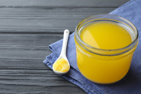 Glass jar and spoon of Ghee butter on gray wooden table, closeup. Space for text