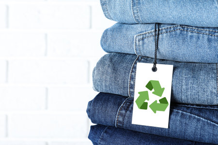 Stack of different jeans with recycling label near white brick wall, closeup. Space for textの写真素材