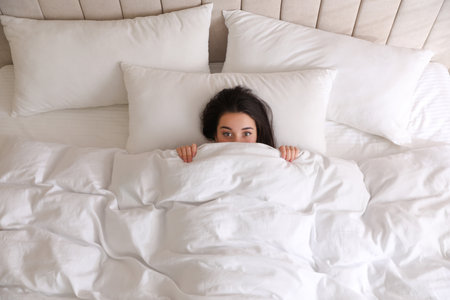 Young woman hiding under warm white blanket in bed, top viewの写真素材