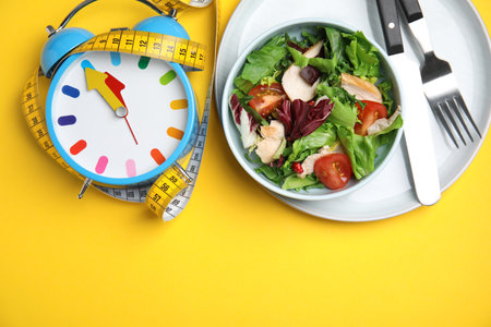 Plate of tasty salad, alarm clock and measuring tape on yellow background, flat lay with space for text. Nutrition regimenの写真素材