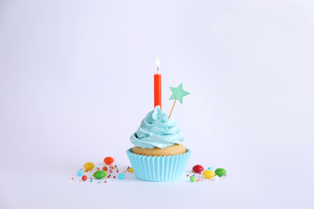 Delicious birthday cupcake with burning candle and candies on light gray backgroundの写真素材
