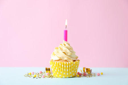 Delicious birthday cupcake with burning candle and sprinkles on white table against pink backgroundの写真素材