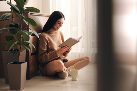 Woman with cup of drink and book sitting on warm floor at home. heating systemの写真素材